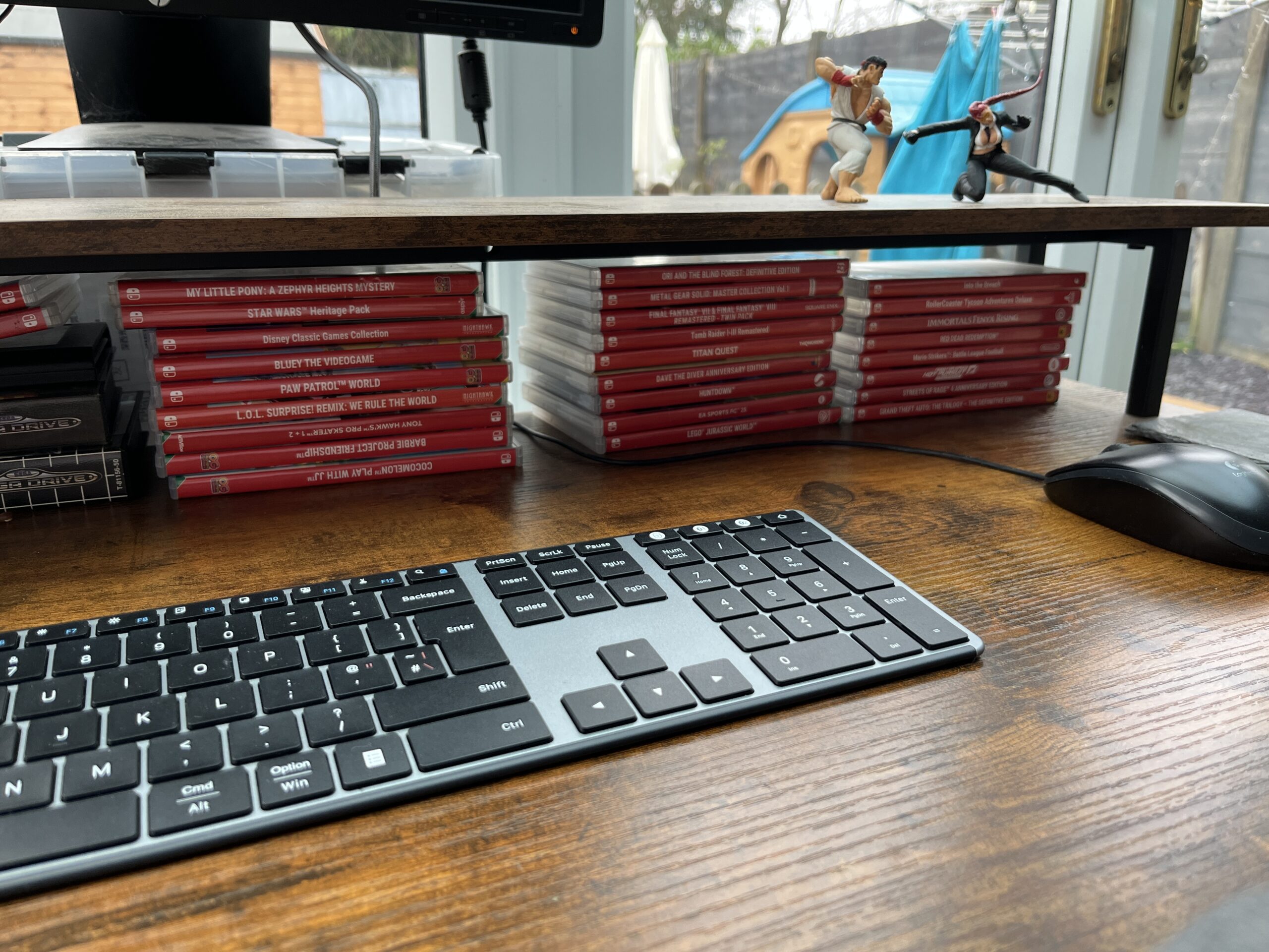 backlog-as-a-library Desk setup with keyboard and mouse, with stacks of Nintendo Switch games stored under a monitor stand.